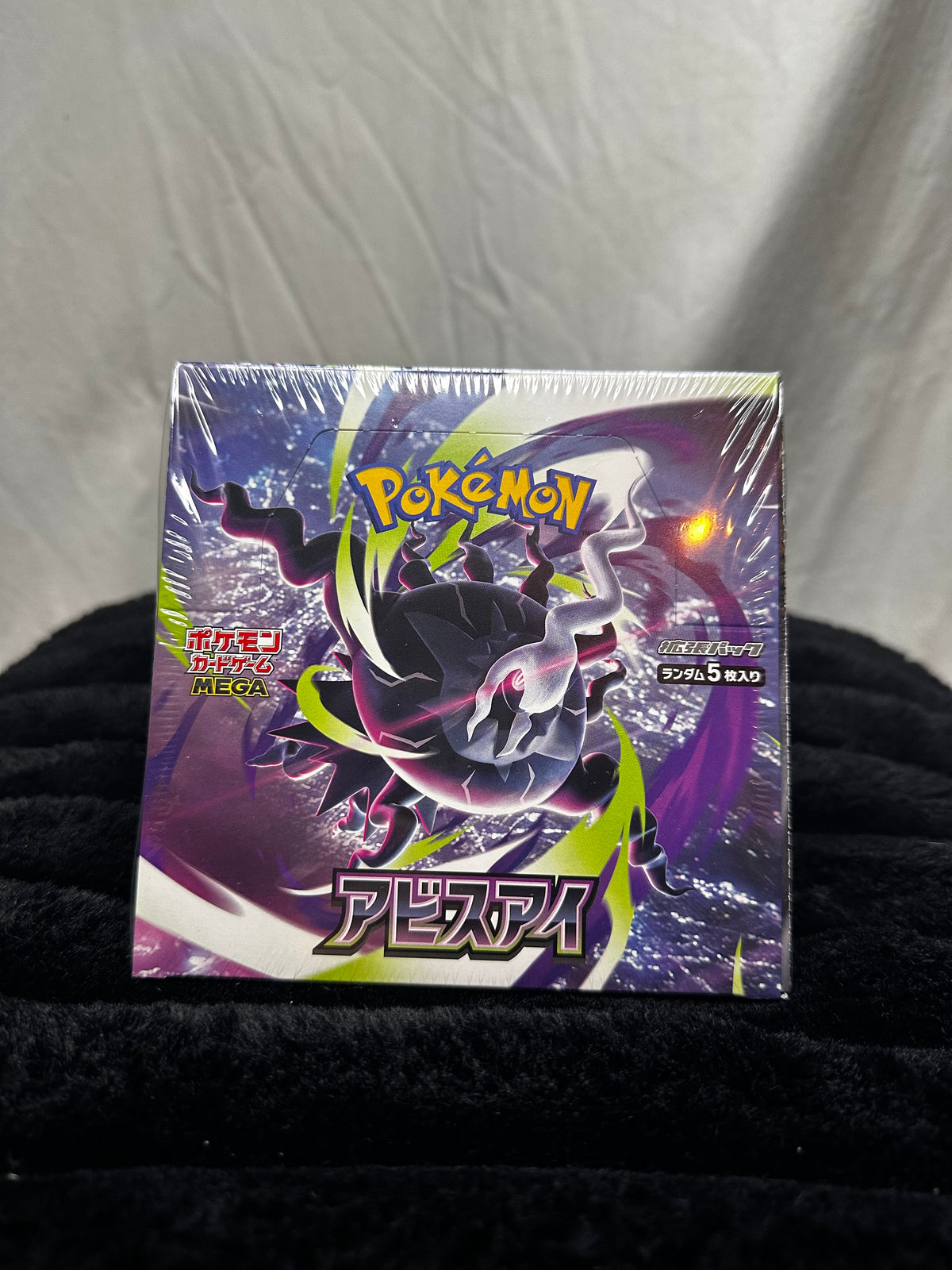 A sealed Pokémon TCG booster pack featuring a large purple and black creature illustration with green accents, photographed on a black fabric surface against a wrinkled light gray backdrop.