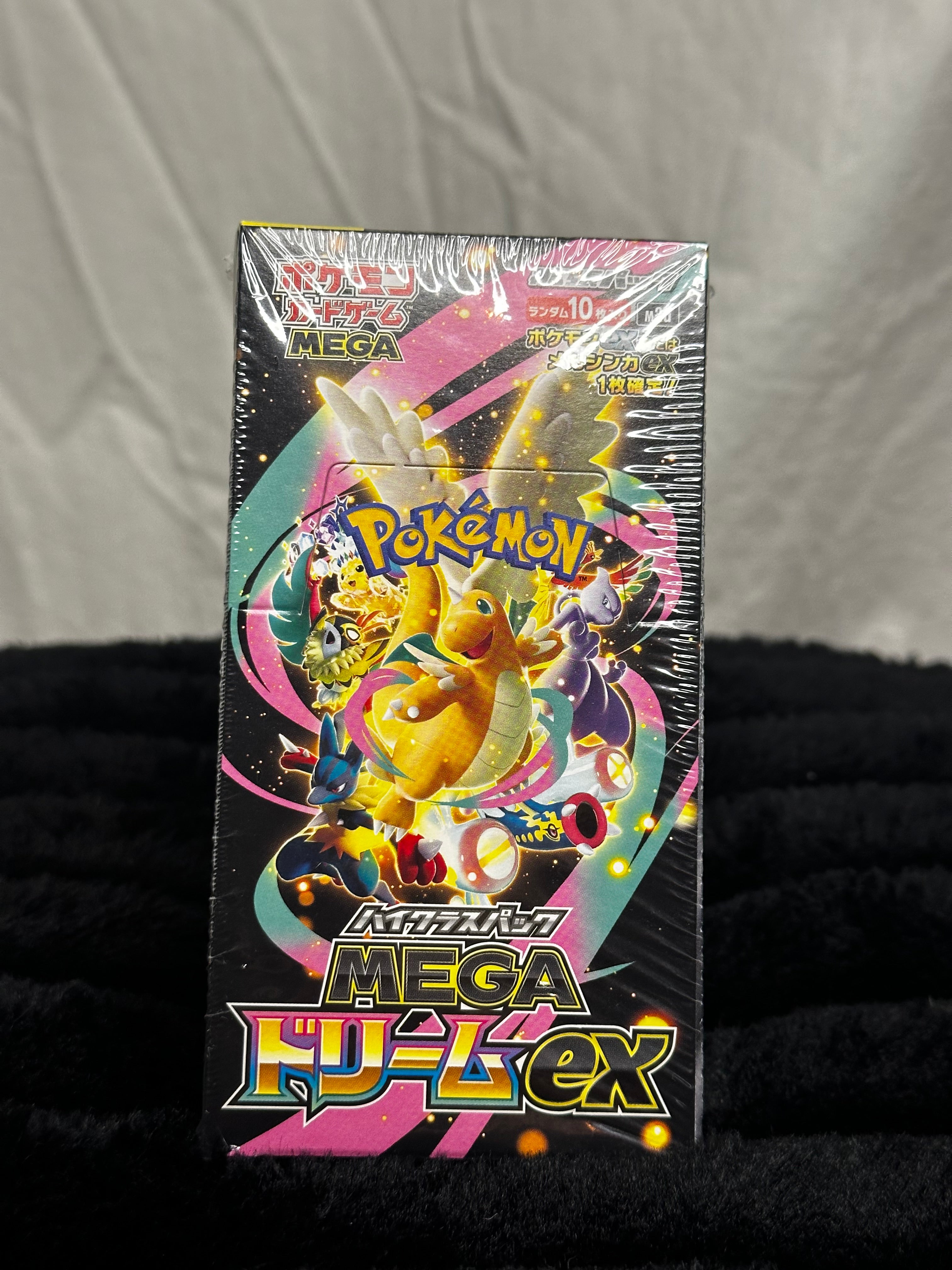 A sealed Pokémon TCG booster pack box for Mega HD-lex, featuring colorful character artwork including Eevee, wrapped in clear plastic, standing against a black fabric background.