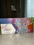 Pokémon TCG box for the Prismatic Evolutions Super-Premium Collection, featuring a holographic rainbow finish and a character illustration, resting on a light-colored surface.